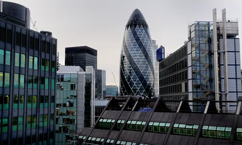 This urban architecture photograph captures a view of city offices in London, United Kingdom, during the late afternoon in the early autumn season. The main subject of the image is the prominent Gherkin building, also known as 30 St Mary Axe, which stands out among the modern glass and steel structures that define the city skyline. The photograph highlights the dense concentration of high-rise office buildings, reflecting the characteristic architectural style of London's financial district, with the iconic Gherkin serving as a recognizable landmark in the heart of the city.